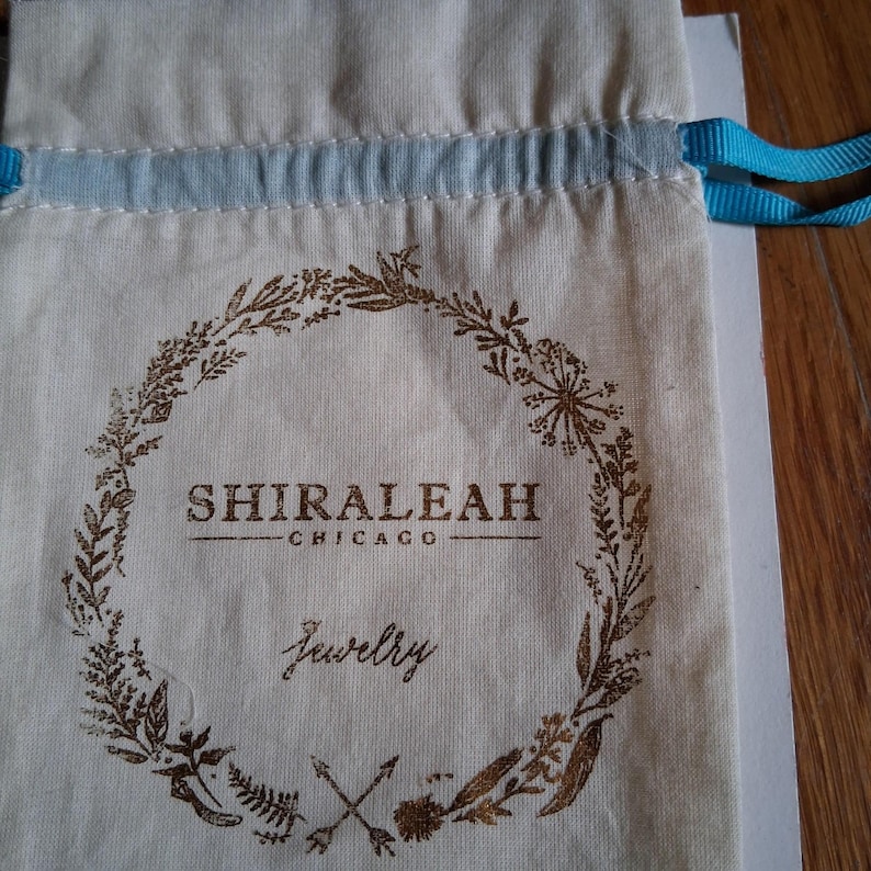 Op de afbeelding: Een cr&egrave;mekleurige sieraden tas met trekkoord en een bruin bloemenkransontwerp. De tas heeft de tekst "SHIRALEAH CHICAGO Jewelry" in bruin. Lichtblauw lint wordt gebruikt voor het trekkoord.