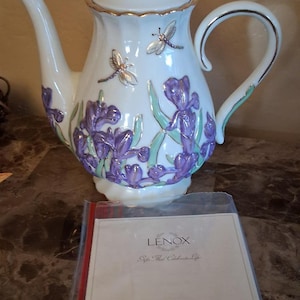 May include: White Lenox teapot with gold trim, a butterfly on the lid, and dragonfly and iris flower designs. The teapot has a curved handle and spout. A document in a plastic sleeve is in front of the teapot.