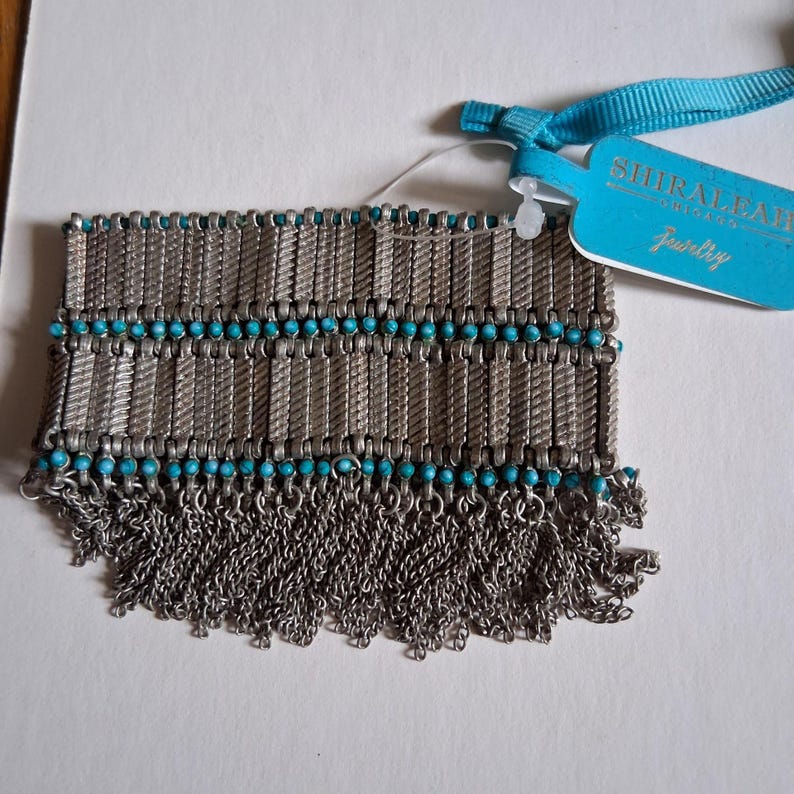 Op de afbeelding: Zilverkleurige armband met turkoois kralen en kettingfranjes. De armband heeft een geweven ontwerp met rijen turkoois kralen. Een blauw label met de tekst "SHIRALEAH ORIGINALS Jewelry" is bevestigd.