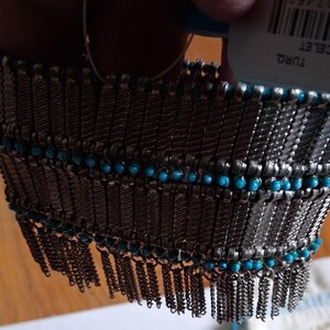 Op de afbeelding: Een zilverkleurige armband met turquoise kralen en bungelende kettingfranjes. De armband heeft een geweven ontwerp met rijen kleine turquoise kralen. Het label luidt "REVERE BRACELET".