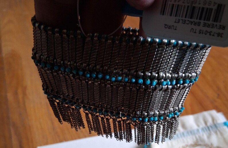 Op de afbeelding: Een zilverkleurige armband met turquoise kralen en bungelende kettingfranjes. De armband heeft een geweven ontwerp met rijen kleine turquoise kralen. Het label luidt "REVERE BRACELET".