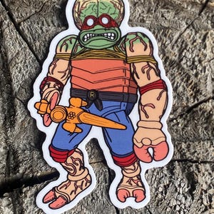 以下が含まれることがあります： 大きな頭と武器を持った緑と赤の漫画のキャラクター。キャラクターは赤と青の服を着て、赤いヘッドバンドをしています。キャラクターは白い背景に立っています。