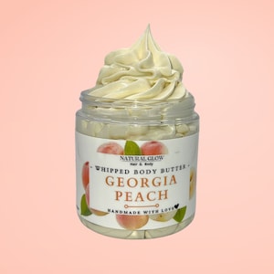 May include: A clear jar of whipped body butter with a peach scent. The jar has a white label with the words "Georgia Peach" and peach illustrations. The whipped butter is a light cream color and is swirled on top of the jar.