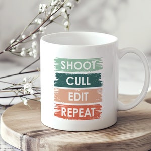 Könnte beinhalten: Weiße Keramiktasse mit dem Text "SHOOT CULL EDIT REPEAT" in grünen, orangefarbenen und roten Farbstrichen. Die Tasse steht auf einem Untersetzer aus Holz.
