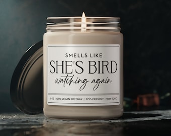 May include: A white candle with a black label that reads "SMELLS LIKE SHE'S BIRD watching again". The candle is in a clear glass jar with a black lid. The candle is 9 oz and made with 100% vegan soy wax. It is eco-friendly and non-toxic.