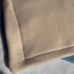 May include: Close-up of a beige blanket with a folded corner. The blanket has a soft, textured surface and a subtle colour variation. The corner is neatly sewn, showing a clean edge and a slight curve.