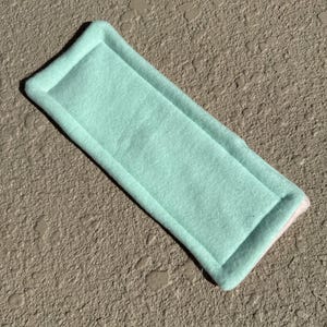 May include: A rectangular, light teal felt pad with a lighter teal centre and a rounded edge. The pad has a stitched border. The item is shown on a textured, neutral-toned surface.