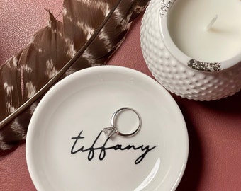 May include: White ceramic ring dish with the word "tiffany" written in black script. A silver ring with a clear stone is sitting in the dish.