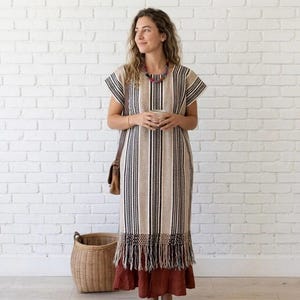 May include: A beige, black, and white striped dress with short sleeves and a fringed hem. The dress is worn over a rust-colored skirt. A brown leather bag is slung over the shoulder. A woven basket sits on the floor.
