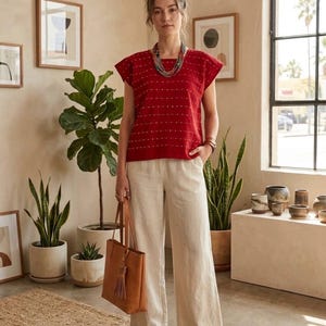 May include: A red short-sleeved top with a white dotted pattern, paired with wide-leg beige trousers. The outfit includes a multi-strand beaded necklace, a tan leather tote bag with a tassel, and tan woven leather sandals on the feet.