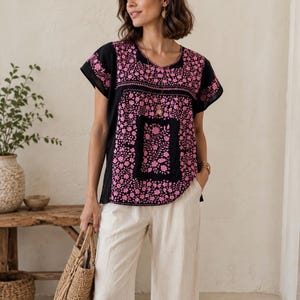 May include: Black short-sleeve top with pink floral embroidery. The top features a square floral design on the front and a round neckline. The model is wearing cream-colored pants and carrying a woven tote bag.