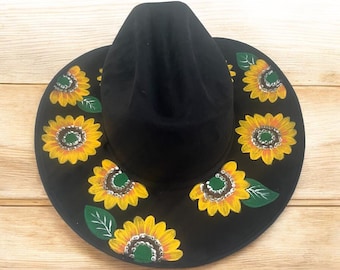 May include: A black cowboy hat with a wide brim decorated with hand-painted sunflowers. The sunflowers have yellow petals, green leaves, and brown centers.