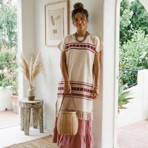 May include: A beige and burgundy patterned dress with a woven basket bag. The dress has short sleeves and a fringed hem. The bag is light brown with a leather handle. The dress is paired with brown sandals.