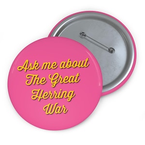 May include: A pink button with the text "Ask me about The Great Herring War" in yellow script. The button has a silver pin back and a shiny silver rim.