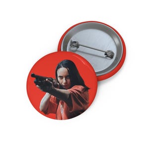 Puede incluir: Un botón circular con alfiler que muestra a una persona de cabello oscuro largo con una camisa naranja apuntando con una escopeta negra. El fondo es de color rojo sólido. La parte posterior es de metal plateado con un mecanismo de alfiler.