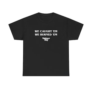 Op de afbeelding: Een zwart T-shirt met korte mouwen en witte tekst op de borst. De tekst luidt WE CAUGHT 'EM WE BURNED 'EM met de woorden WIDOW'S BAY in een kleiner lettertype direct onder de twee hoofdregels met tekst gedrukt.