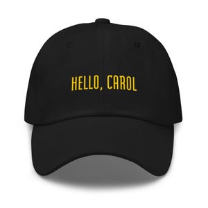 Op de afbeelding: Zwarte baseballpet met de tekst "HELLO, CAROL" in gele letters. De pet heeft een gebogen klep en een ronde kroon. De hoed is gemaakt van een zacht materiaal.