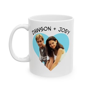 May include: White ceramic mug featuring a heart-shaped image of two people with the text "DAWSON + JOEY" in black. The image inside the heart is of a man and a woman. The mug has a white handle.