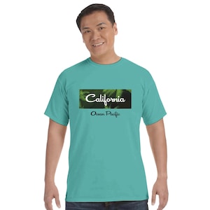 May include: A turquoise t-shirt with the word "California" in white script inside a black rectangle. Below the word "California" is the text "Ocean Pacific" in a smaller font. The shirt is made of a soft material.
