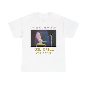 May include: White t-shirt featuring a graphic of a singer with pink hair playing a keyboard under a spotlight. The text "TABITHA JOHANSSON" is above the graphic, and "OIL SPILL WORLD TOUR" is below.