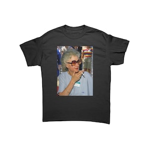 May include: A black and white photo of a woman wearing sunglasses and a light blue shirt. She is holding a cigarette in her right hand and is looking to the right. The photo is printed on a black t-shirt.