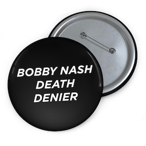 Peut inclure: Deux badges ronds noirs avec le texte "BOBBY NASH DEATH DENIER" en blanc. Un badge est face visible, l'autre est légèrement derrière, montrant l'arrière-plan de l'épingle argentée.