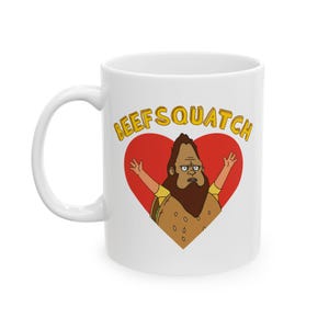 Könnte beinhalten: Weiße Keramiktasse mit einer Cartoon-Illustration eines Bigfoot-ähnlichen Charakters in einem roten Herzen. Die Figur hat einen braunen Bart und erhobene Arme. Die Worte "BEEFSQUATCH" stehen in gelben Buchstaben darüber.