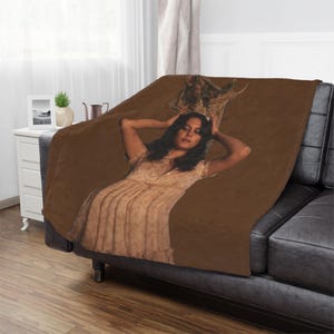 Puede incluir: Una manta marrón sobre un sofá de cuero negro. La manta tiene una imagen impresa de una mujer con un vestido. La mujer está apoyando la cabeza en sus manos. La manta tiene una textura suave y afelpada. El sofá está en un salón.