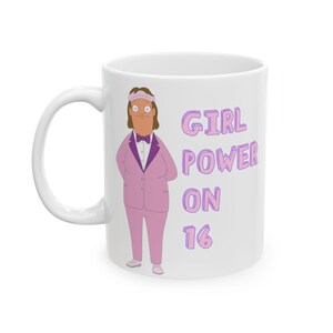 May include: White ceramic mug featuring a cartoon character in a pink suit with a bow tie. The mug has the text "GIRL POWER ON 16" in purple. The handle is white.