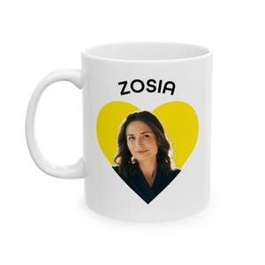 May include: White ceramic mug featuring the name "ZOSIA" in black letters above a yellow heart-shaped design. The heart contains a portrait of a woman. The mug has a curved handle.