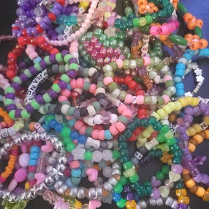 May include: A collection of colorful beaded bracelets in various sizes and designs. The bracelets feature a mix of colors, including pink, green, purple, and orange, with some spelling out words. Some bracelets have heart and star-shaped beads.