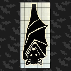 May include: Black silhouette of a bat hanging upside down with its wings spread out. The bat is facing the viewer. The image is on a white background with a grid pattern.