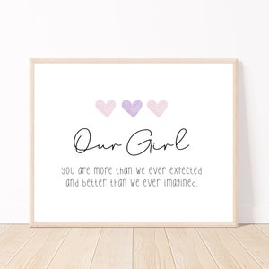 May include: A printable wall art design with a white background and a black cursive font that reads "Our Girl" with three watercolour hearts in shades of pink and purple above the text. The text below the hearts reads "you are more than we ever expected and better than we ever imagined."