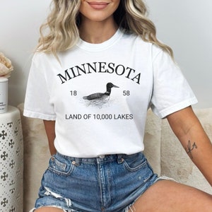 May include: White t-shirt with black text that reads "Minnesota" in an arc above a black and white illustration of a loon. Below the loon is the text "18" and "58". Below the loon is the text "Land of 10,000 Lakes".