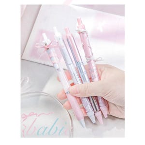 May include: A hand holding five pastel-coloured pens with decorative bows. The pens are in shades of pink, white, and silver. The word "babi" is visible in the lower left corner.