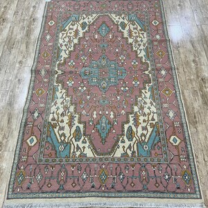 Puede incluir: Una alfombra rectangular con un detallado patrón geométrico en tonos rosa, crema, turquesa y dorado. La alfombra tiene un diseño de medallón central y un borde con flecos. Las dimensiones son 2,69 metros por 1,60 metros.