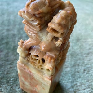 Könnte beinhalten: Eine geschnitzte Steinskulptur einer mythischen Kreatur, möglicherweise eines Wächterlöwen, in Beige- und Cremetönen. Die Skulptur steht auf einem rechteckigen Sockel mit roten und cremefarbenen Marmorierungen. Die Schnitzerei weist komplizierte Details und ein stilisiertes Design auf.