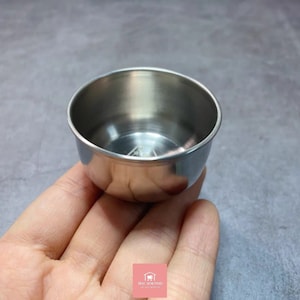 Pode incluir: Uma pequena tigela de aço inoxidável com formato arredondado. A tigela é segurada em uma mão, e o interior reflete a luz ambiente. A tigela é provavelmente usada para servir molhos ou pequenas porções.