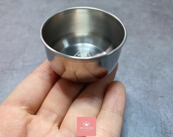 May include: A small, stainless steel bowl with a rounded shape. The bowl is held in a hand, and the interior reflects the surrounding light. The bowl is likely used for serving sauces or small portions.
