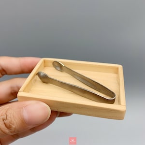 Puede incluir: Una pequeña bandeja de madera clara con un par de pinzas de color plateado en su interior. Las pinzas están diseñadas con extremos redondeados. La bandeja es rectangular con un borde elevado.