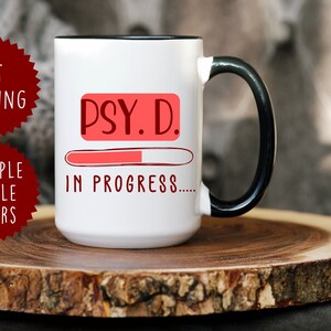 Könnte beinhalten: Weiße Keramik-Kaffeetasse mit schwarzem Rand und Henkel. Die Tasse hat einen roten Fortschrittsbalken mit dem Text "PSY. D. IN PROGRESS..." darauf gedruckt.