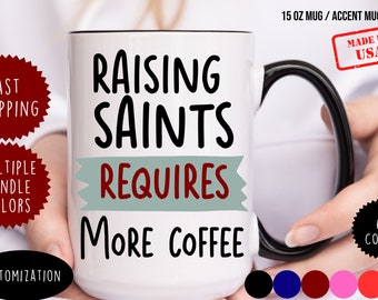 May include: White ceramic mug with a black handle and rim. The mug has black text that reads "Raising Saints Requires More Coffee".