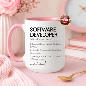 May include: White ceramic mug with a pink handle and interior. The mug features the text "SOFTWARE DEVELOPER" and a definition, including the phrase "99 bugs in the code. Fix one. 127 bugs remain." A gold-colored spoon rests nearby.
