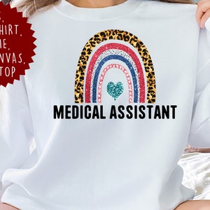 May include: A white long-sleeve sweatshirt featuring a central graphic of a rainbow with leopard print, red glitter, and blue glitter stripes. A teal glitter heart sits below the rainbow, with the text MEDICAL ASSISTANT printed in black below.
