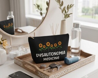 Pode incluir: Uma bolsa preta com zíper apresentando um gráfico de cinco girassóis e o texto branco "DYSAUTONOMIA MEDICINE". A bolsa está posicionada em uma bandeja de madeira ao lado de uma garrafa de água transparente, um inalador azul, chaves e moedas.
