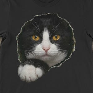 May include: Black t-shirt featuring a cat's face with yellow eyes and white markings, appearing to peek through a torn hole. The shirt has a crew neck and short sleeves. The word "BLACK" is printed at the bottom. A phone with the Etsy logo is visible.