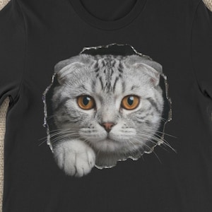 May include: Black t-shirt featuring a Scottish Fold cat design. The cat's face and paw appear to be emerging from a torn hole in the shirt. The cat has gray and white fur with orange eyes. The Etsy logo is visible on a smartphone.