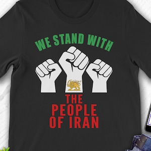 Puede incluir: Camiseta negra con el texto "WE STAND WITH THE PEOPLE OF IRAN" en verde y rojo. El diseño presenta tres puños blancos, uno con un emblema dorado. También se ve un teléfono con el logotipo de Etsy y unos vaqueros.