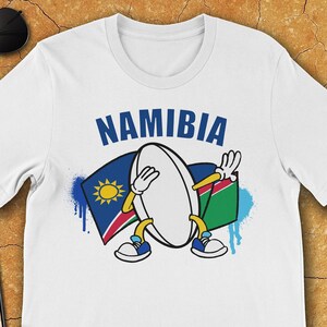 Peut inclure: T-shirt blanc avec un ballon de rugby dessin animé portant un drapeau namibien bleu et vert. Le texte "NAMIBIA" est au-dessus du dessin animé.