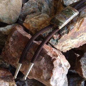 May include: A bolo tie with a rectangular, patterned stone pendant in shades of green, brown, and cream. The tie features a dark brown braided cord with silver-colored tips. The bolo tie rests on a bed of various-sized rocks.
